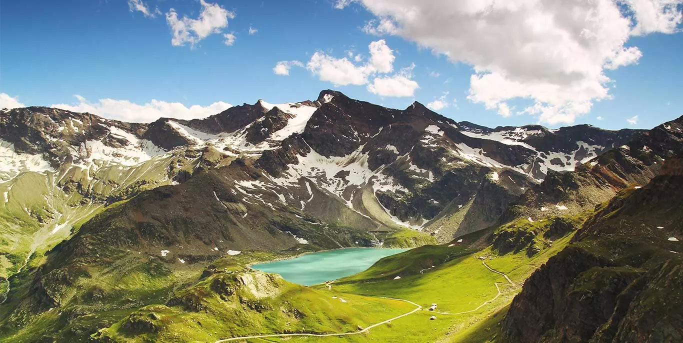 Landschaftsbild leicht schneebedeckte Berge unter leicht bewölktem Himmel mit grünen Wiesen und blaugrünem Bergsee
