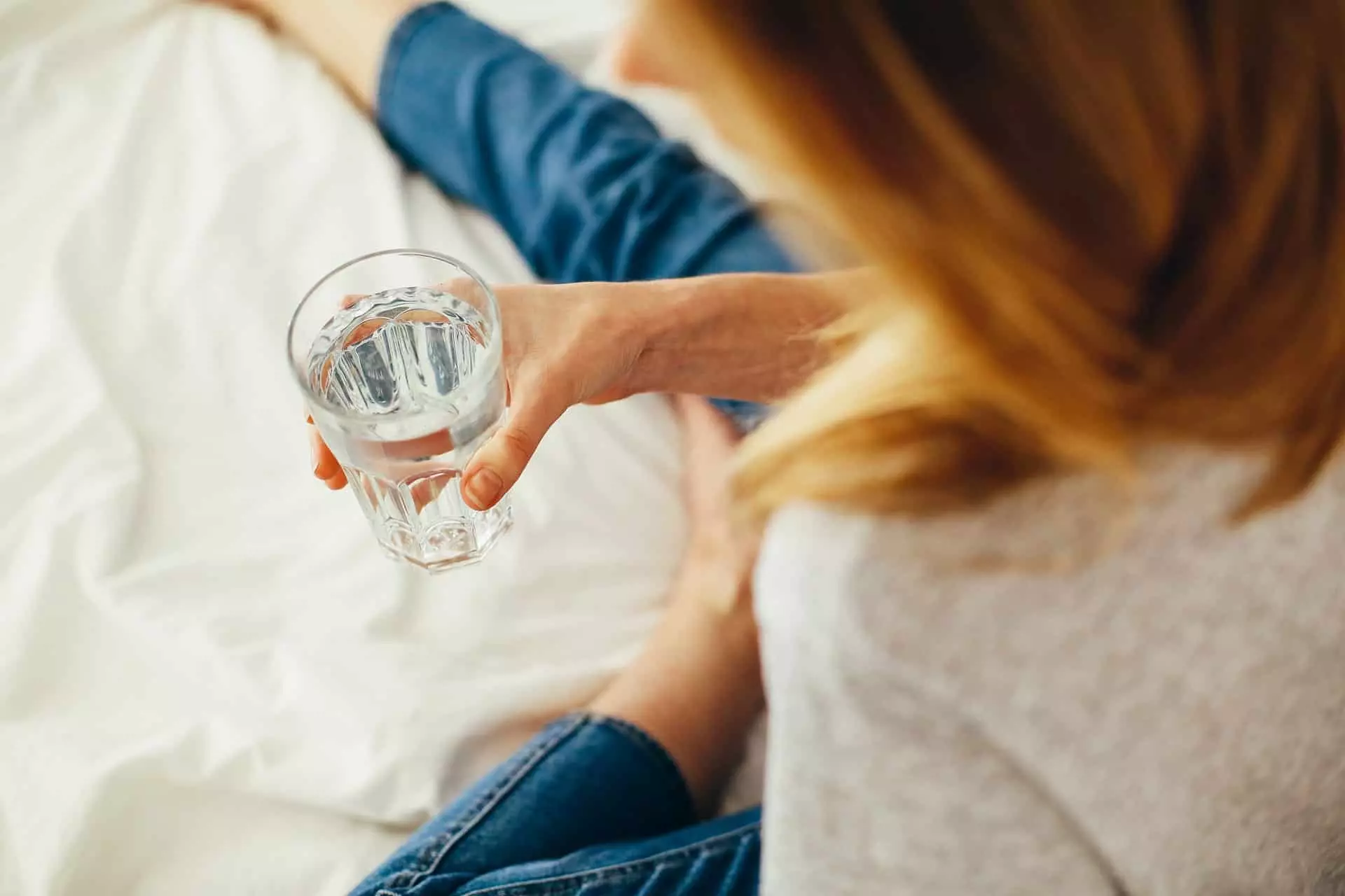 Person mit Wasserglas in der Hand, sitzend auf einem Bett